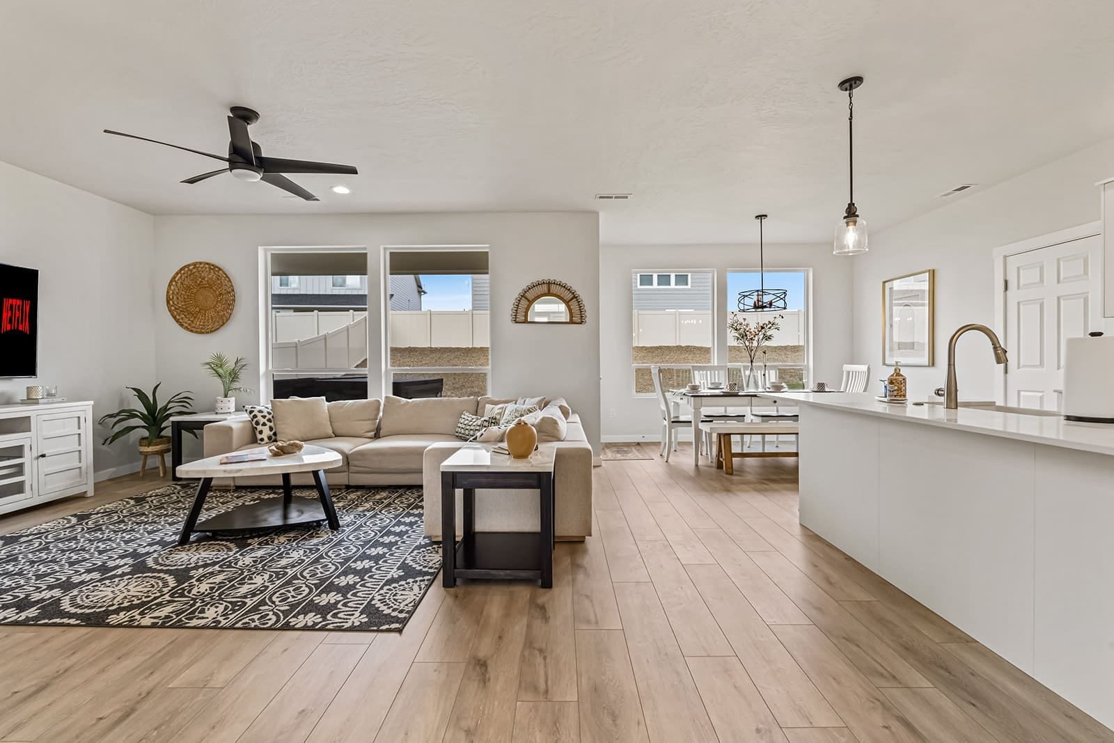 A modern living room with a sofa, coffee table, and a TV. There is a dining area with a table and chairs. The room has wooden flooring and white walls.