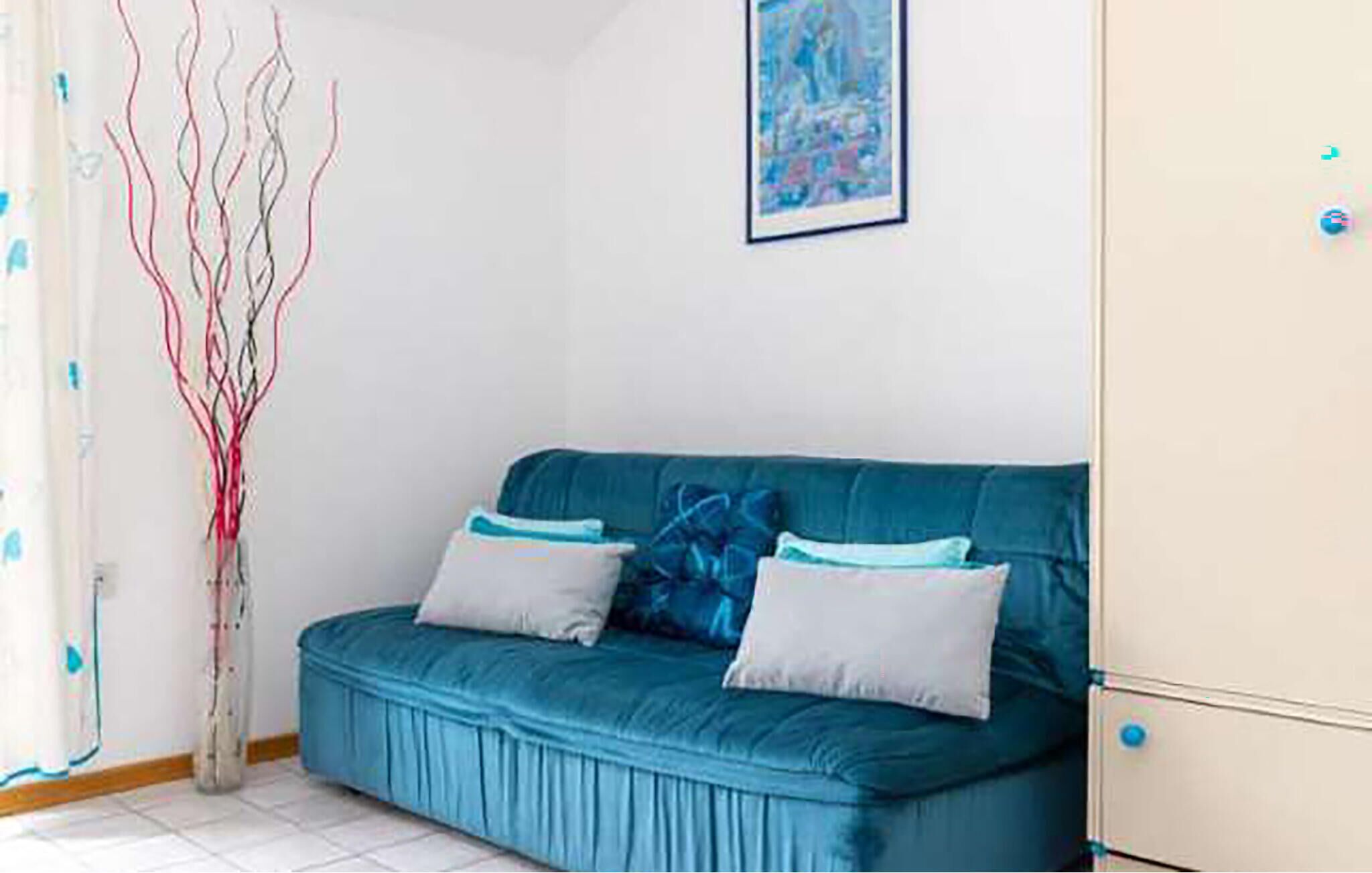 Une pièce avec un canapé bleu, deux coussins, un vase avec des branches, un tableau au mur et un meuble.