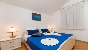 A neatly made bed with blue bedding, white pillows, and a decorative towel arrangement. There is a bedside table with a lamp, a framed picture on the wall, and a window with white curtains. - Pet friendly apartment in Bol with lake view (Bol)