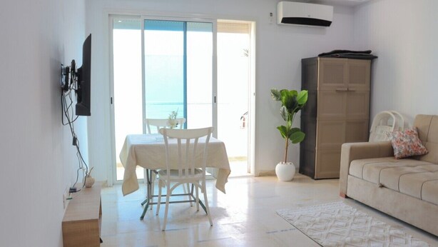 A living room with a sofa, a dining table with chairs, a potted plant, and a sliding glass door leading to a balcony.