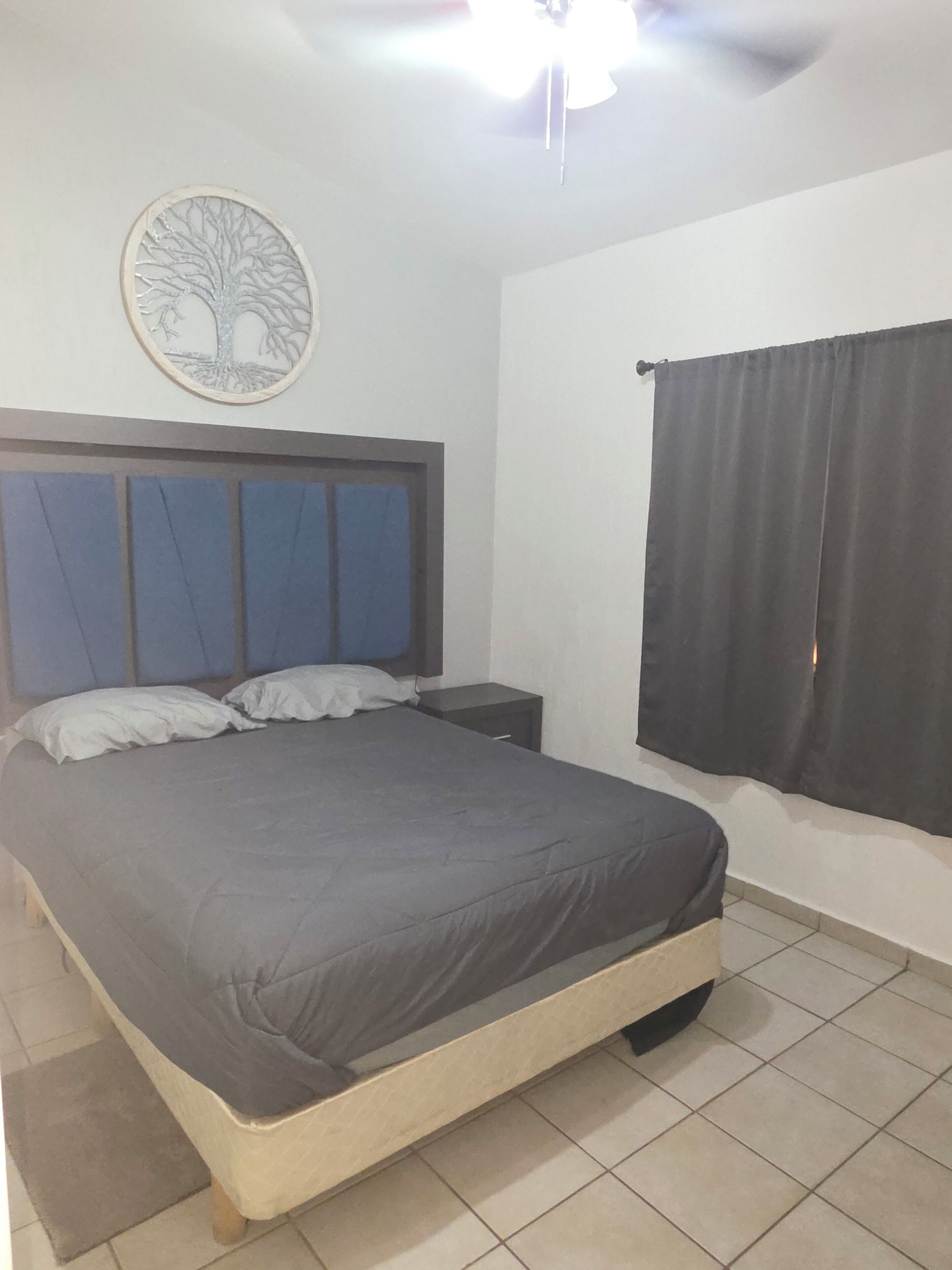 Une chambre à coucher comprenant un lit, une table de chevet, une œuvre d’art accrochée au mur et une fenêtre avec des rideaux.
