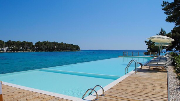 Ein klares, blaues Schwimmbecken mit Holzterrasse und Liegestühlen, mit Blick auf ein ruhiges Meer und ein von Bäumen gesäumtes Ufer.