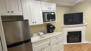 A modern kitchen with white cabinetry, a stainless steel refrigerator, a microwave, a coffee maker, and a flat-screen TV above a fireplace. -  Awesome Ocean Front Vacation Condo on the Cape in MA (Dennis)
