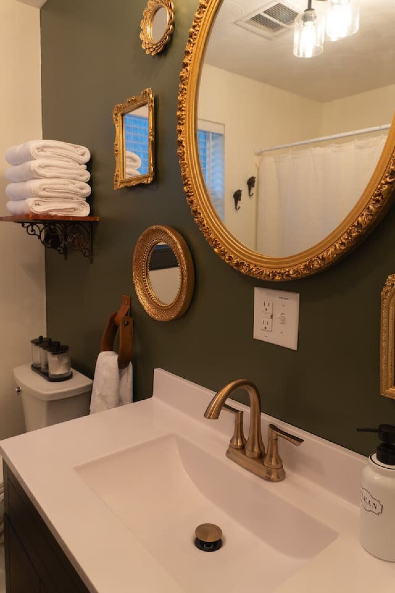 A bathroom with a white sink, gold faucets, and a large mirror with a gold frame. There are towels stacked on a shelf and a toilet visible in the background.