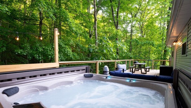 A hot tub with seating and a table, surrounded by a wooden deck and trees. - The Mountain Magnolia *Hot tub on private deck* (Inez)