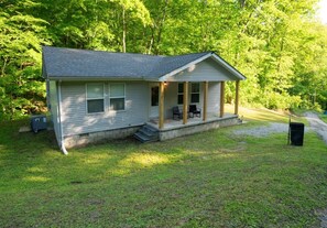 A single-story cabin with a porch, a small front yard, and a wooded backdrop.
