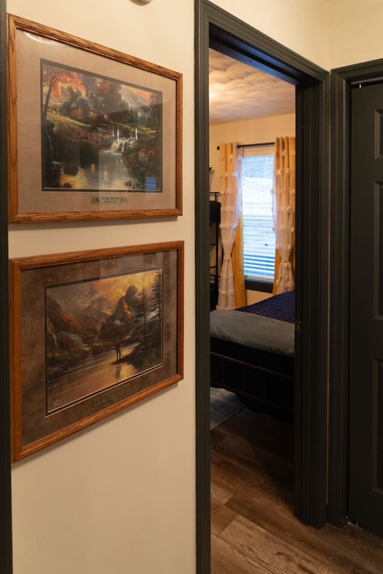 Ein Flur mit zwei gerahmten Bildern an der Wand, einem Bett mit blauer Decke und einer Tür zu einem Zimmer mit Fenster und Vorhängen.