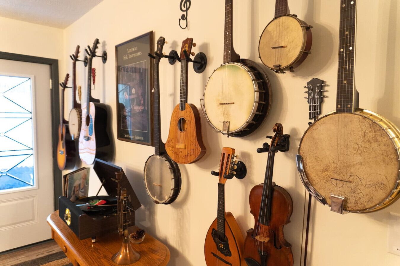 A room with a wall-mounted guitar and banjo collection, a framed certificate, and a small table with a guitar case.