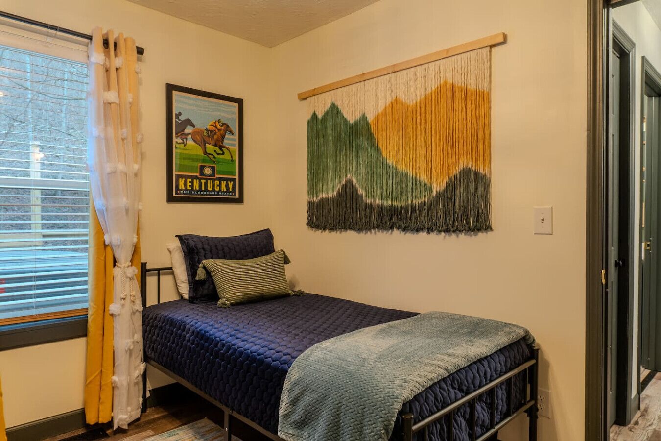 A single bed with a blue quilt, a framed Kentucky state poster, and a mountain landscape tapestry on the wall.