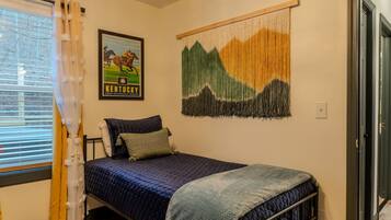 A single bed with a blue quilt, a framed Kentucky state poster, and a mountain landscape tapestry on the wall.
