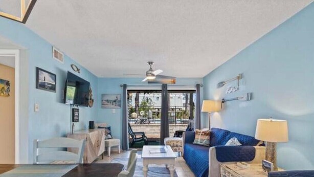 A room with a blue wall, a ceiling fan, a sofa, a coffee table, and a TV mounted on the wall. - Tranquil 2-bedroom condo with Wolf bay views (Orange Beach)