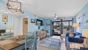 A living room with a dining table, chairs, a sofa, and a TV. There is a door leading to a balcony with a view. - Tranquil 2-bedroom condo with Wolf bay views (Orange Beach)