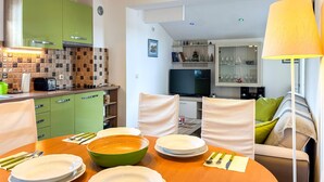 Un comedor con una mesa de madera dispuesta para cuatro, armarios verdes, un televisor y una lámpara.