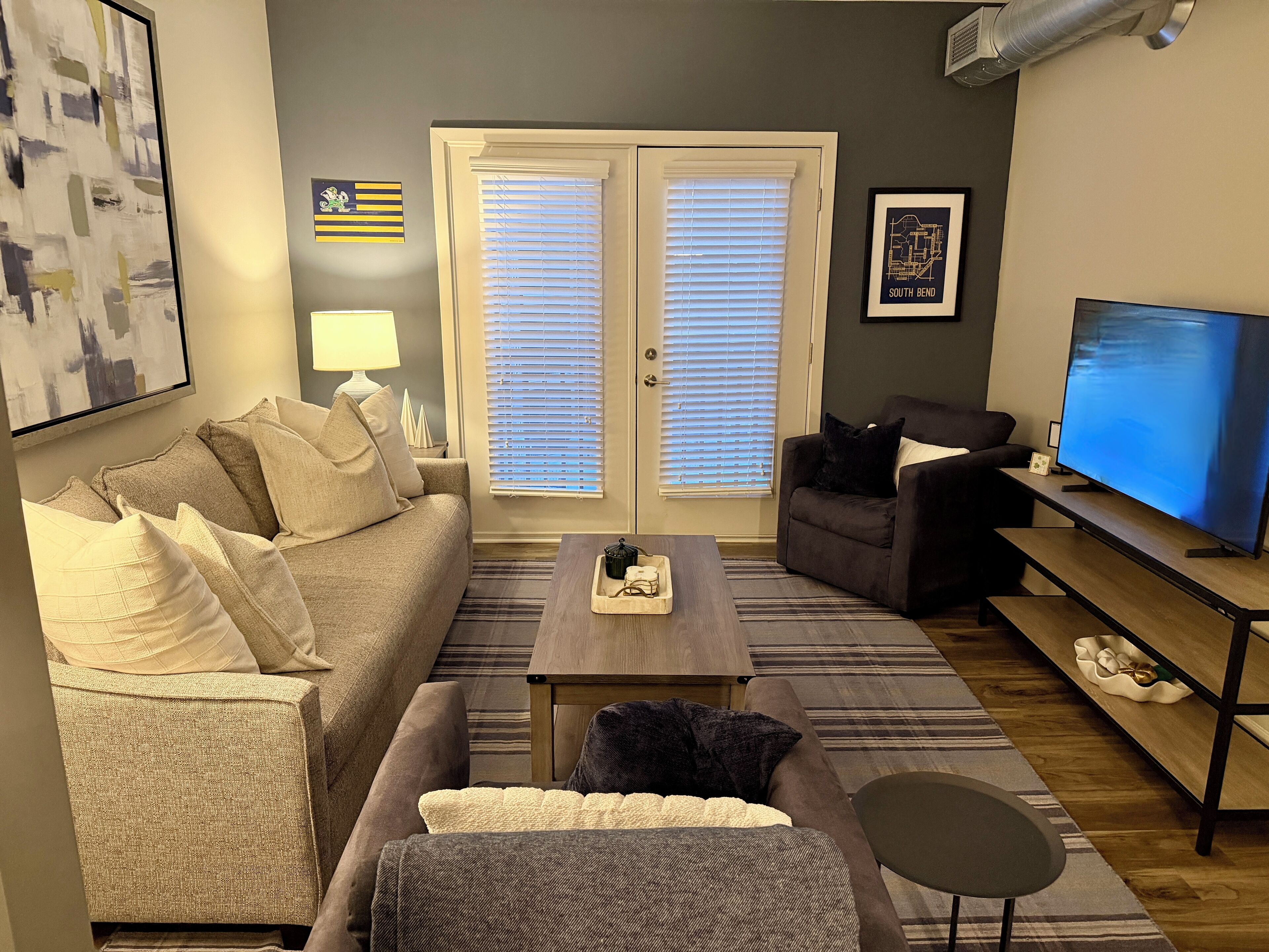 En stue med en beige sofa, en mørk lænestol, et sofabord og et fjernsyn på et stativ.
