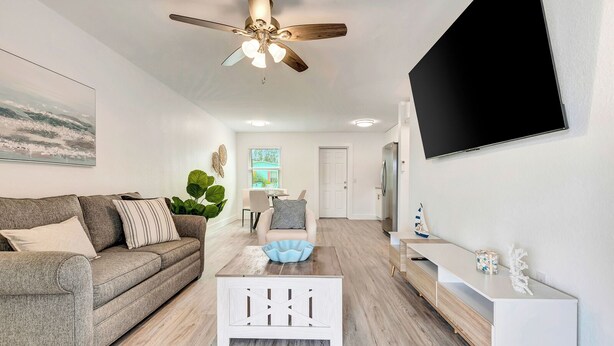 A modern living room with a grey sofa, a white coffee table, a ceiling fan, a TV mounted on the wall, and a door leading to another room. - The Cozy Cove II 3 min to the beach (Bradenton)