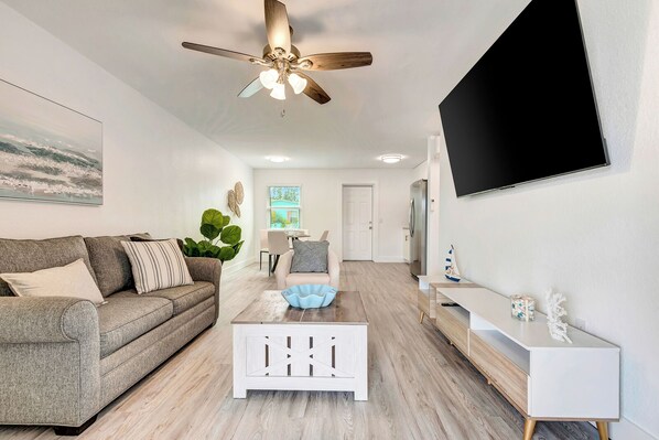 A modern living room with a grey sofa, a white coffee table, a ceiling fan, a TV mounted on the wall, and a door leading to another room.