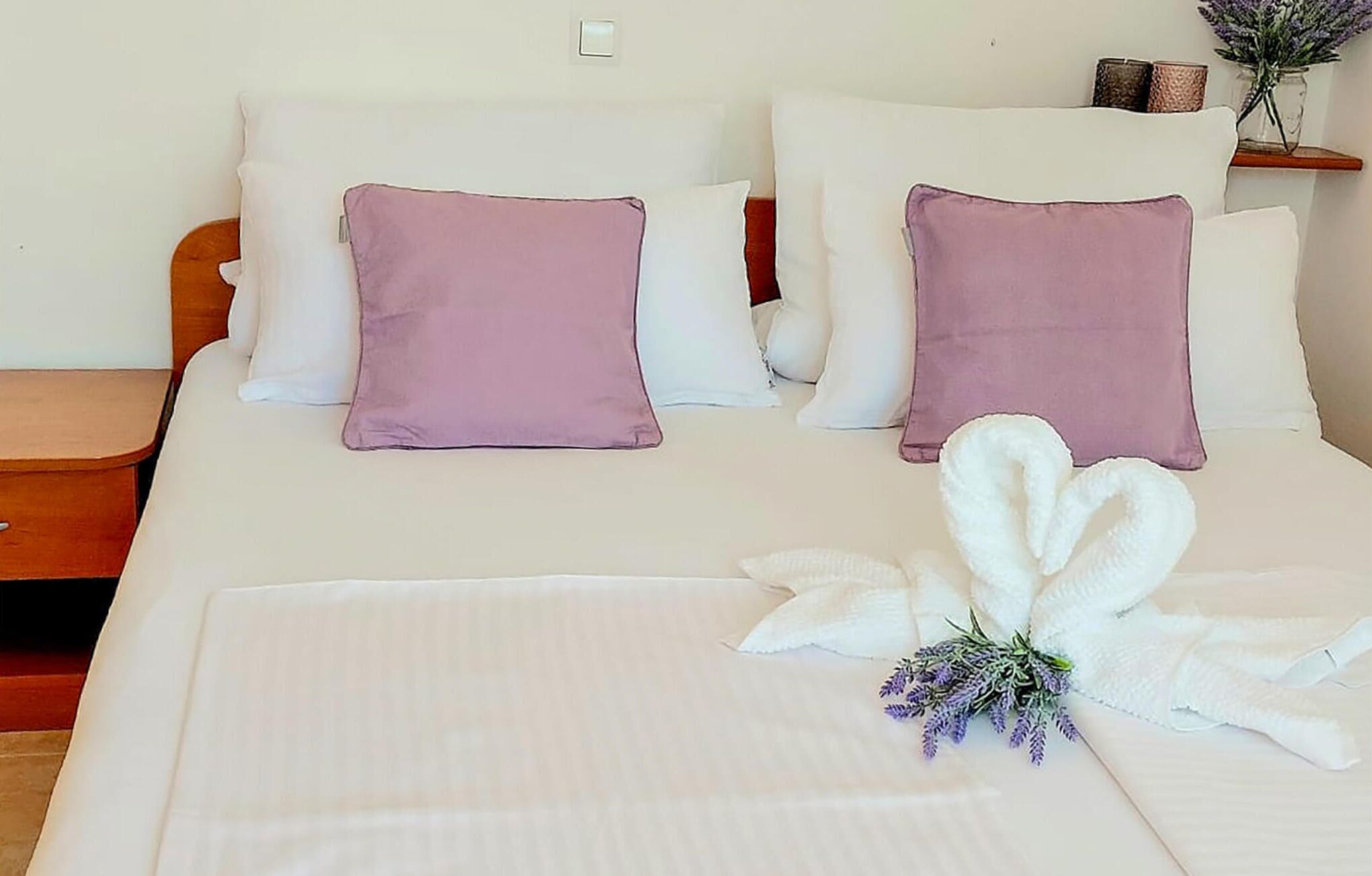 Ein ordentlich gemachtes Bett mit weißen und lila Kissen, ein Handtuch in Weiß und ein kleiner Lavendelstrauß.