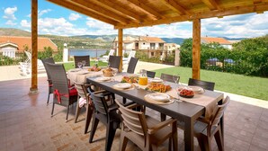 Un espace de restauration en plein air agrémenté d’une pergola en bois, une table dressée pour six personnes avec des assiettes, des verres et une carafe de jus d’orange.