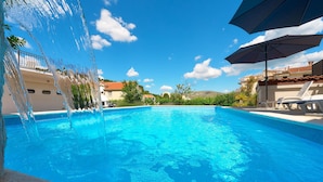 Une piscine d’un bleu éclatant, agrémentée d’une fontaine, entourée de chaises longues et de parasols, avec des maisons et de la verdure en arrière-plan.