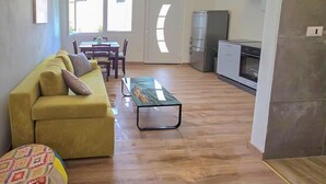 Een woonkamer met een gele bank, een salontafel en een kleurrijk voetenbankje. Er is een eethoek met een tafel en stoelen, en een keuken met een koelkast en oven.