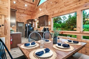 A wooden cabin interior with a dining area set for four, kitchen with modern appliances, and a large window overlooking greenery. - A Spectacular View! *Hot Tub*Games*Pool*Dollywood! (Sevierville)