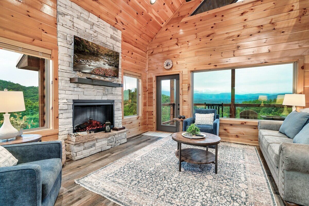L’intérieur d’une cabane en rondins, avec une cheminée en pierre, des murs en bois, une grande fenêtre et un coin salon confortable.