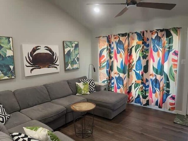 A modern living room with a grey sectional sofa, a round wooden coffee table, and colorful tropical-themed curtains. - Tropical Heaven - Unit 1600 - Island Dunes Condos (Port Aransas)