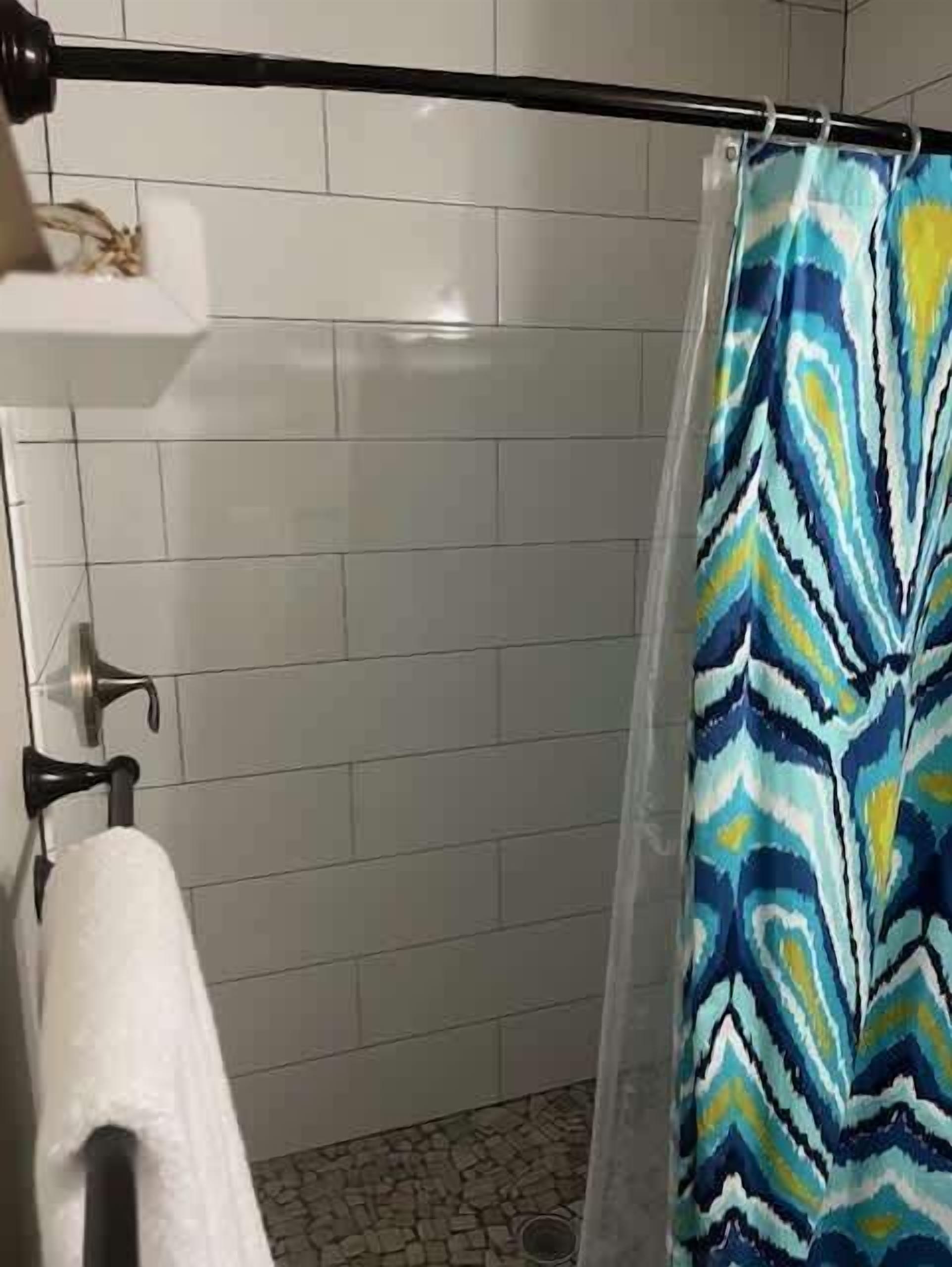 Une salle de bain avec des murs carrelés de carreaux blancs, un rideau de douche à motifs bleus et jaunes, et une serviette blanche suspendue à un porte-serviettes.