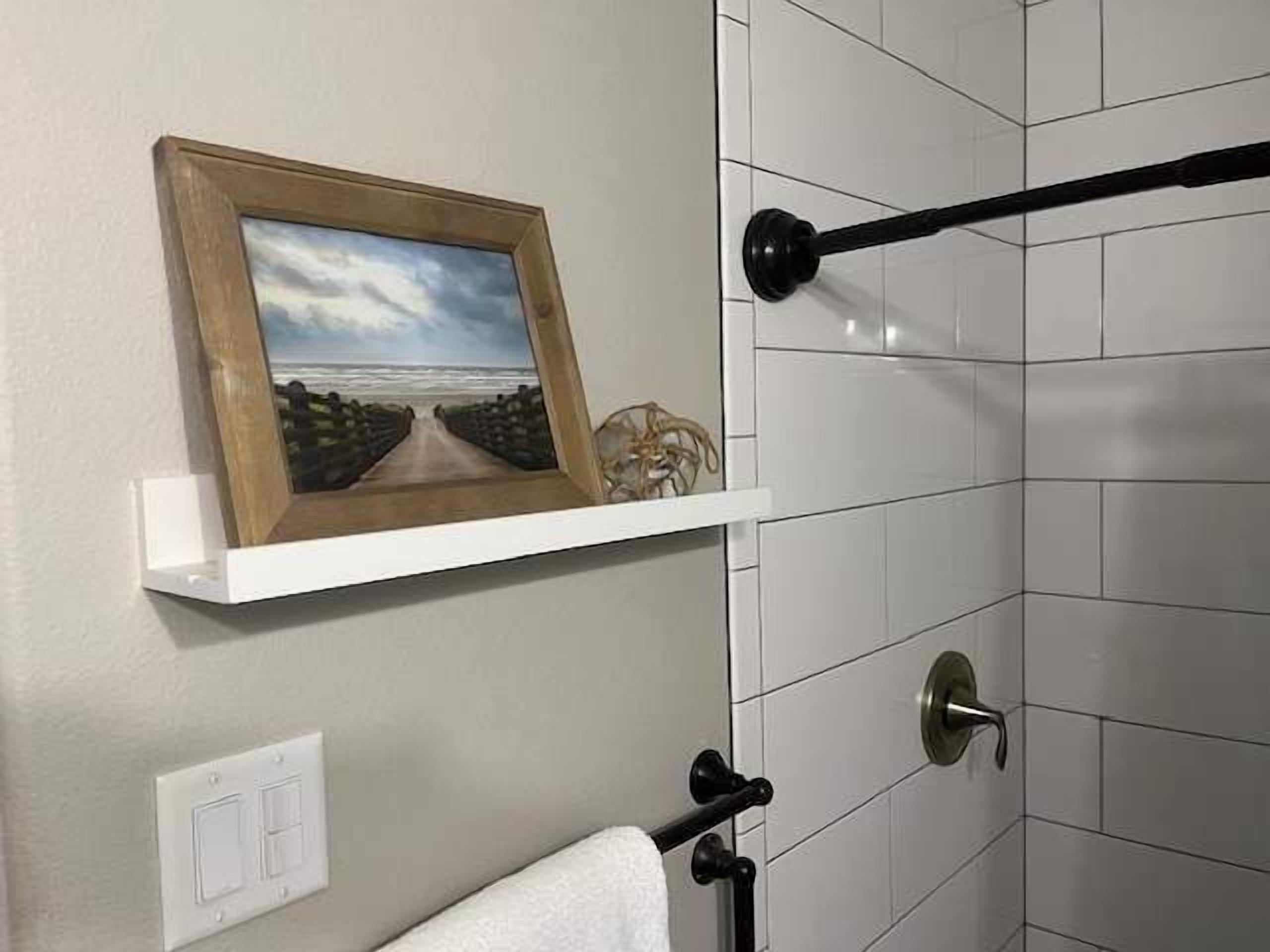 A bathroom with white tiles, a framed picture on the wall, a towel rack, and a shower curtain rod.