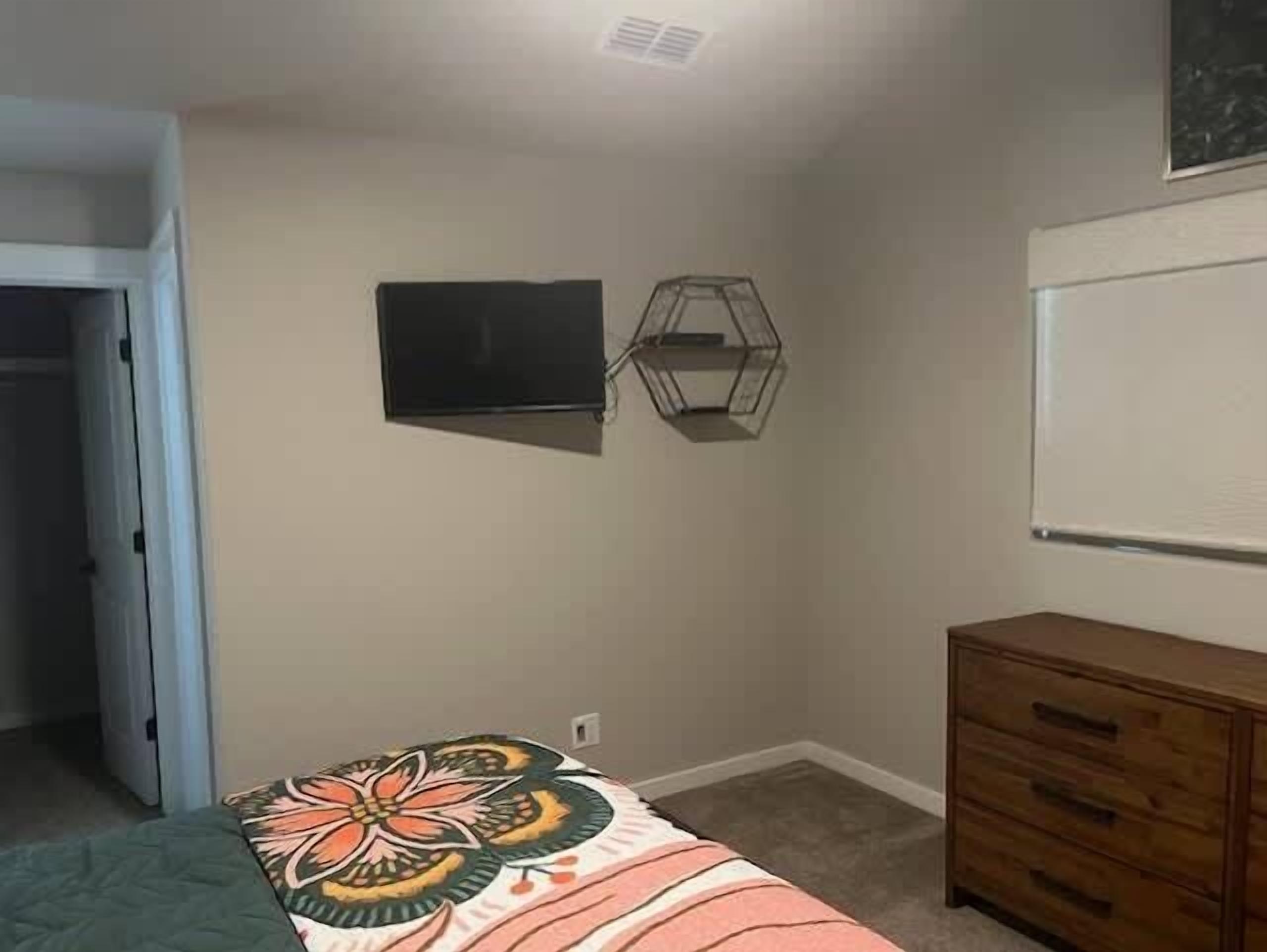A bedroom with a bed, a dresser, a television mounted on the wall, and a window.