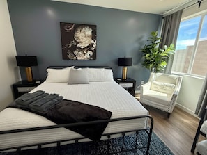 A modern bedroom with a bed, two bedside tables, a chair, a large window, and a wall-mounted artwork.
