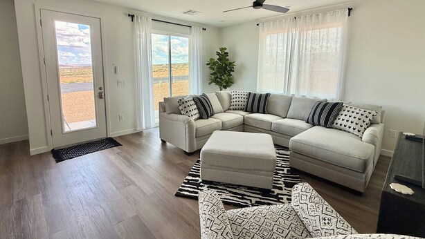 Ein großzügiges Wohnzimmer mit einer breiten Eckcouch, einem kleinen Hocker und Blick auf eine Wüstenlandschaft durch eine Schiebetür.