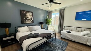 Ein modernes Hotelzimmer mit einem Bett, einem Einzelbett, einem Schreibtisch, einem Stuhl, einem Deckenventilator, einem Fernseher und einem Fenster mit Vorhängen.
