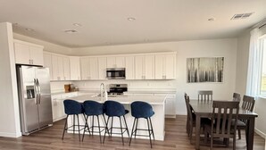 Eine moderne Küche mit weißen Schränken, einem Edelstahlkühlschrank, einer Mikrowelle und einem Spülbecken. Vier blaue Barhocker und ein Holztisch mit Stühlen ergänzen das Bild.