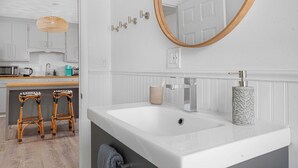 A modern bathroom with a white sink, a round mirror, and a wooden stool. - Warren 1BR Retreat + Spa | Walk to Dining & Water (Warren)