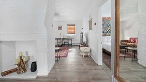Um quarto amplo com piso de madeira, paredes de tijolos brancos, uma cama, um espaço para refeições e um espelho refletindo parte do quarto.