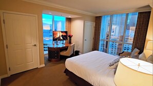 Ein Hotelzimmer mit einem großen Bett, einem Schreibtisch mit Fernseher und Blick auf eine Stadtlandschaft durch große Fenster.