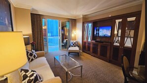 Ein Hotelzimmer mit Sofa, Ohrensessel, Glastisch und Fernseher auf einer Holzkommode.
