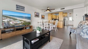 A living room with a large flat-screen TV, a dining area with white chairs, a kitchen with wooden cabinets, and a ceiling fan. - Beachfront Getaway in St. Augustine (St. Augustine)