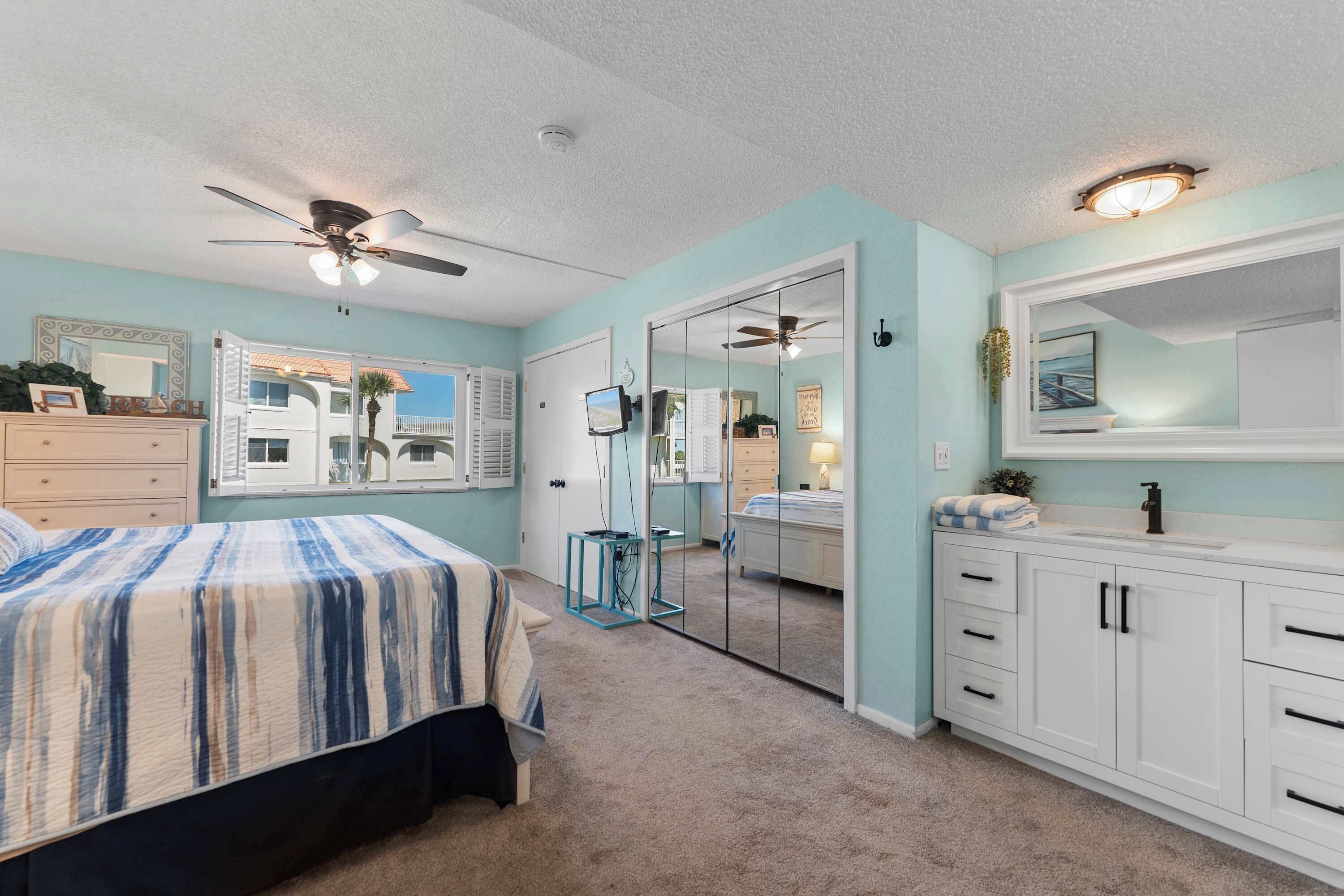 A bedroom with a bed, a ceiling fan, a mirror, a dresser, and a window.