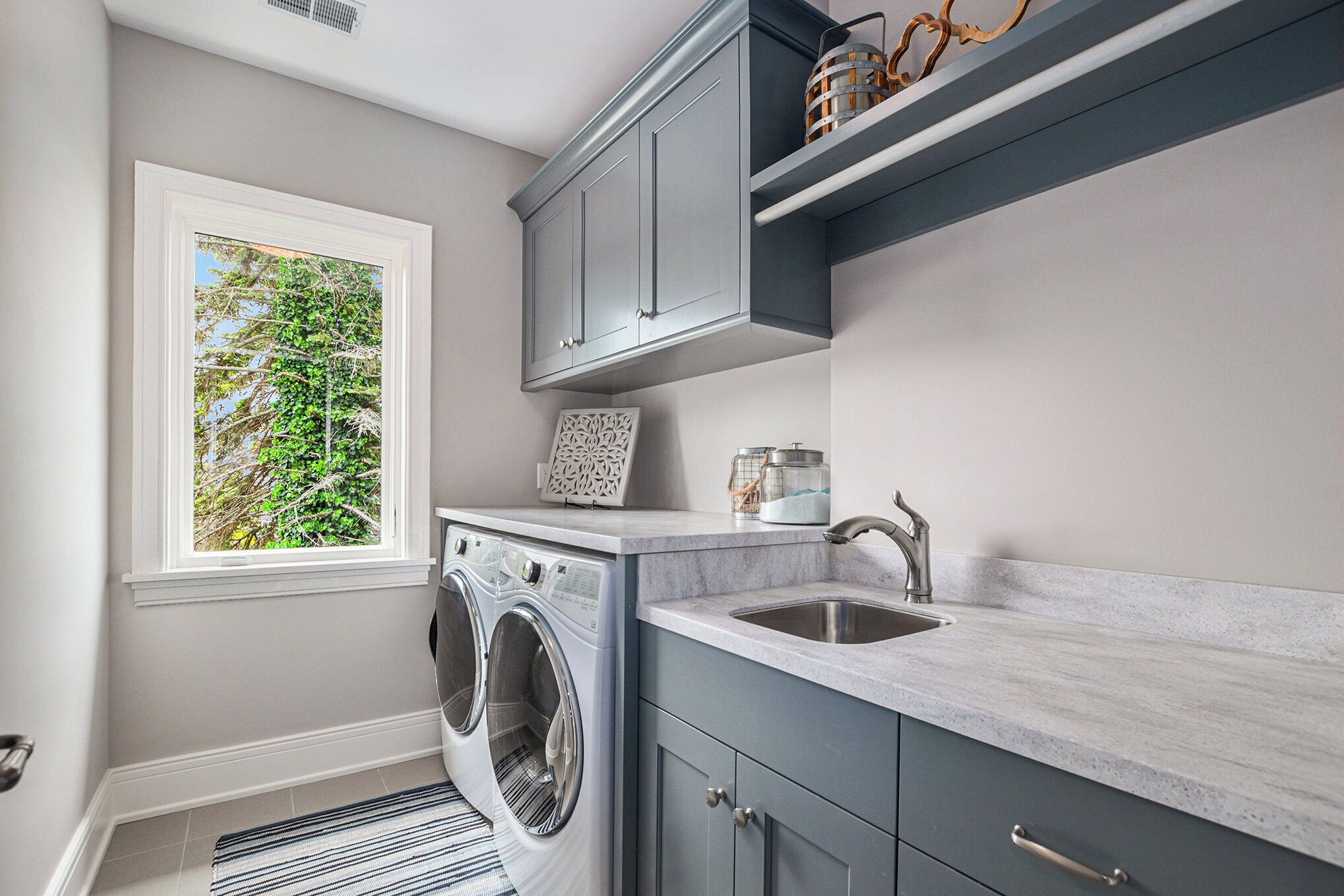 A modern laundry room with a built-in washer and dryer, a window with a view of greenery, and a countertop with a sink.