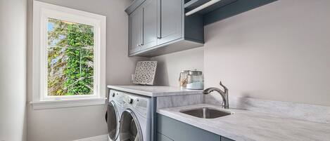 A modern laundry room with a built-in washer and dryer, a window with a view of greenery, and a countertop with a sink.