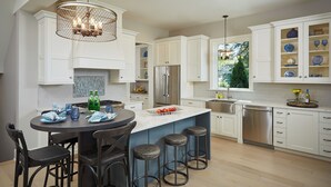 Une cuisine moderne avec des meubles blancs, un îlot central doté de tabourets de bar, des appareils en acier inoxydable et un lustre décoratif.