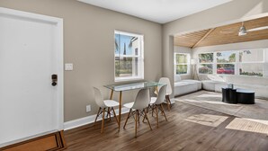 Un salon moderne avec une table à manger en verre, des chaises blanches, une table basse noire et une grande fenêtre avec des stores.