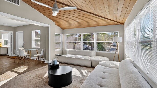 Un salon moderne avec un plafond en bois, des canapés blancs, un coin repas avec une table et des chaises, et de grandes fenêtres équipées de stores.