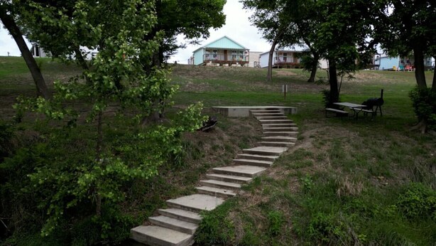 Una escalera de hormigón que asciende por una colina cubierta de hierba, rodeada de árboles y un pequeño cuerpo de agua.