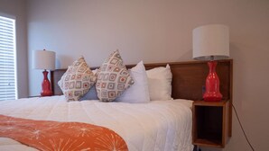Una cama bien hecha con un edredón blanco y naranja, dos almohadas estampadas y dos lámparas de noche rojas.