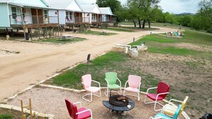 Un camping con sillas de colores dispuestas alrededor de un brasero central, un sendero de tierra que conduce a un edificio con porche y una zona verde con árboles al fondo.