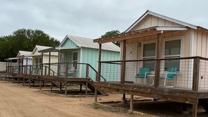 A row of small, single-story cabins with wooden decks and railings, each featuring a single chair and a small table. - Kerrville Standalone Cabin near Downtown & River (Kerrville)
