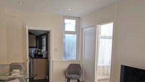 Uma sala de estar moderna com um sofá, uma televisão, uma mesa de vidro e uma poltrona. Cozinha visível por uma porta aberta.